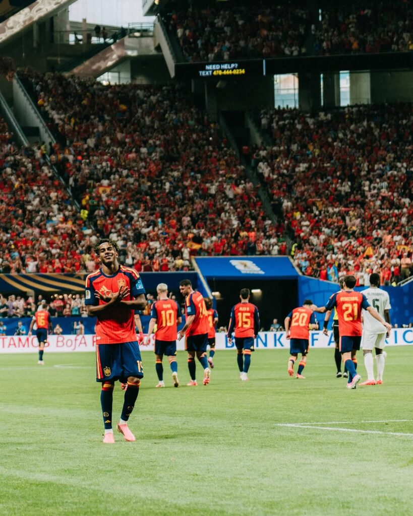 Im here vamos españa gracias a todos por el apoyo siempre con dios🤞🏾🤎🇪🇸 1 catrawarta Soccer player in a red and blue kit smiles at the camera on a green pitch with a full stadium behind him